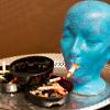 Blue mannequin head smoking a cigarette surrounded by ash trays