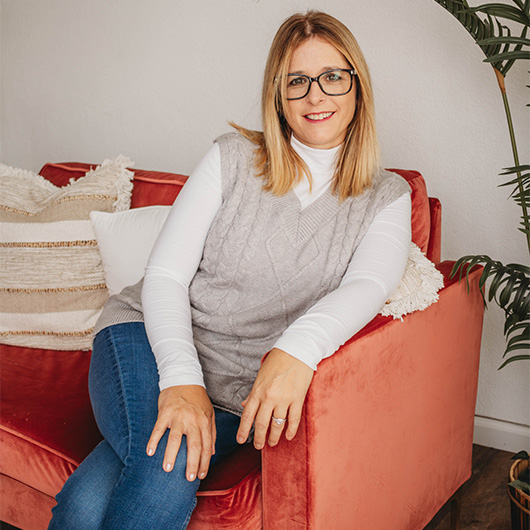 Woman sitting on a red couch, wearing jeans and a grey sweater, and glasses