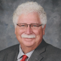 A smiling Ron Sullivan, wearing framed glasses, a grey suit jacket over a grey and white checkered shirt and red tie.