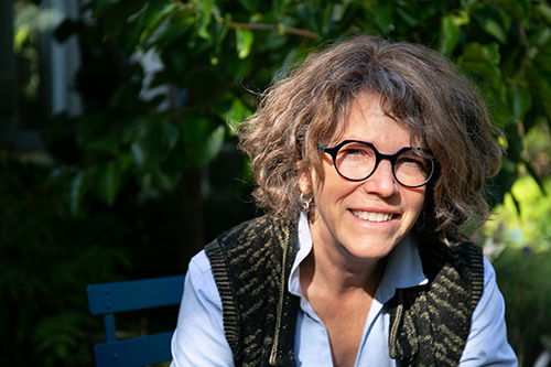 Woman wearing black glasses sits on a blue bench in front of green trees