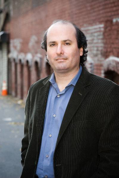 David Grann, wearing a blue button-up shirt and black blazer, standing in front of a brick wall.