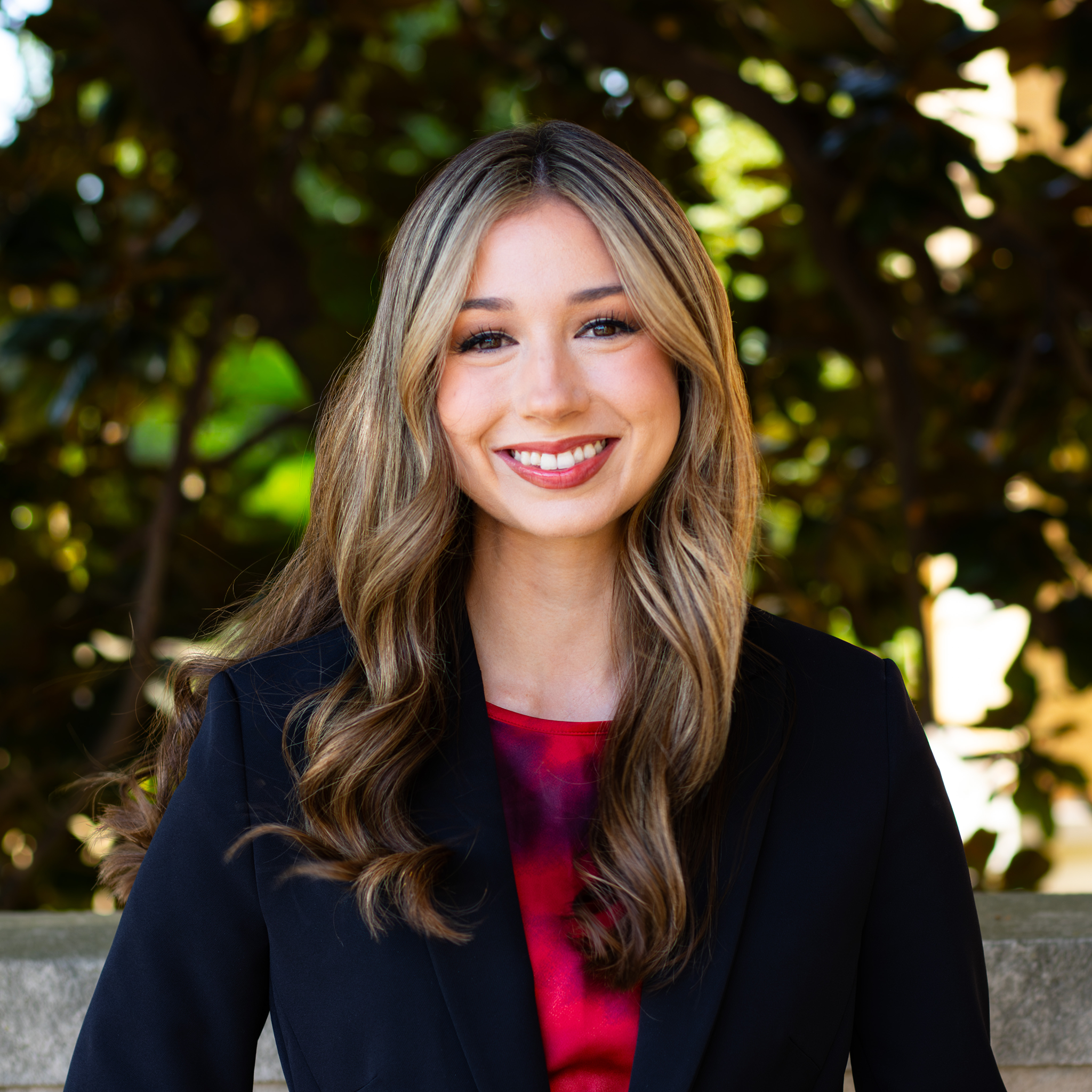 A woman standing in a black suit and red shirt smiling at the camera
