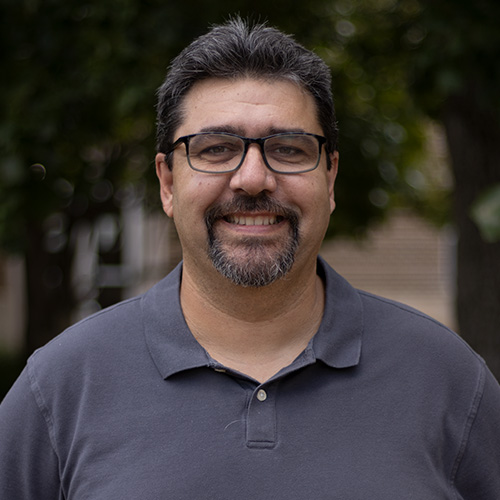 Smiling man with black hair, a goatee and black glasses, wearing a medium grey polo shirt