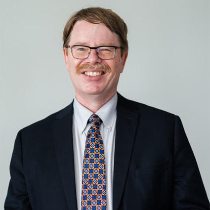Smiling man with light brown hair, glasses, and a moustache, wearing a charcoal suit and a copper tie with blue squares and diamonds on it