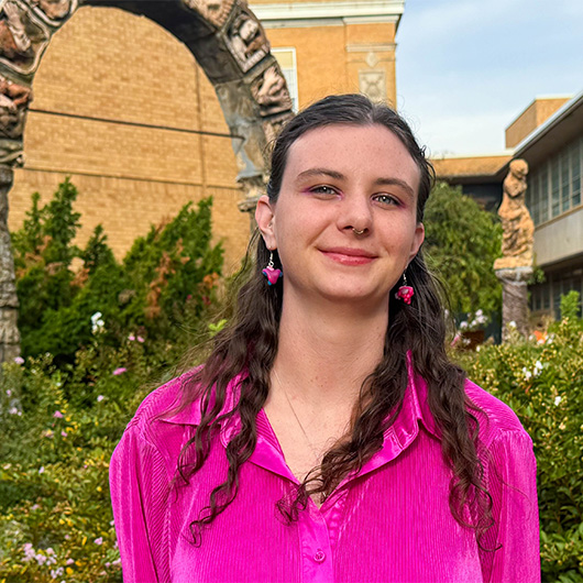 woman in pink shirt and earrings standing in front of archway and foliage