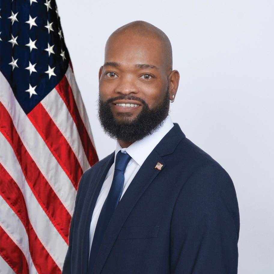 A man in a navy suit standing in front of American flag and smiling
