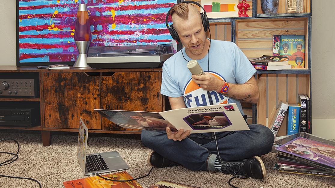 A photo of Michael Bendure sitting in front of a microphone with piles of vinyl funk records on either side