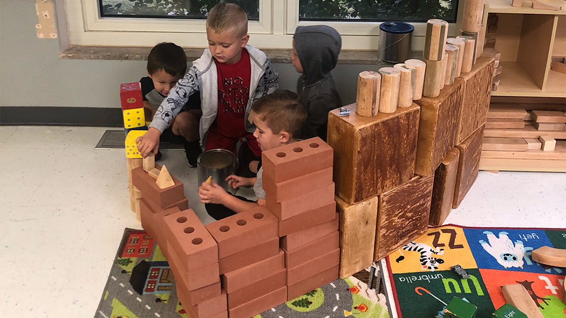 A photo of four young children playing in a fort made of foam blocks in the shape of logs and bricks