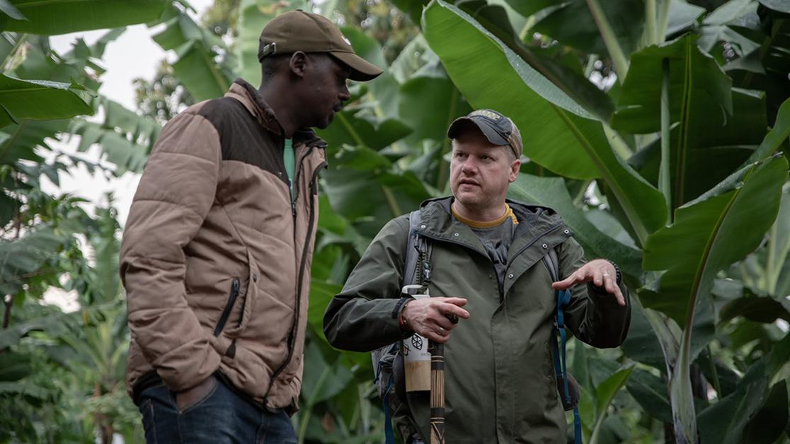 A photo of Cody Lorrance speaking with a Ugandan coffee supplier in a forested setting