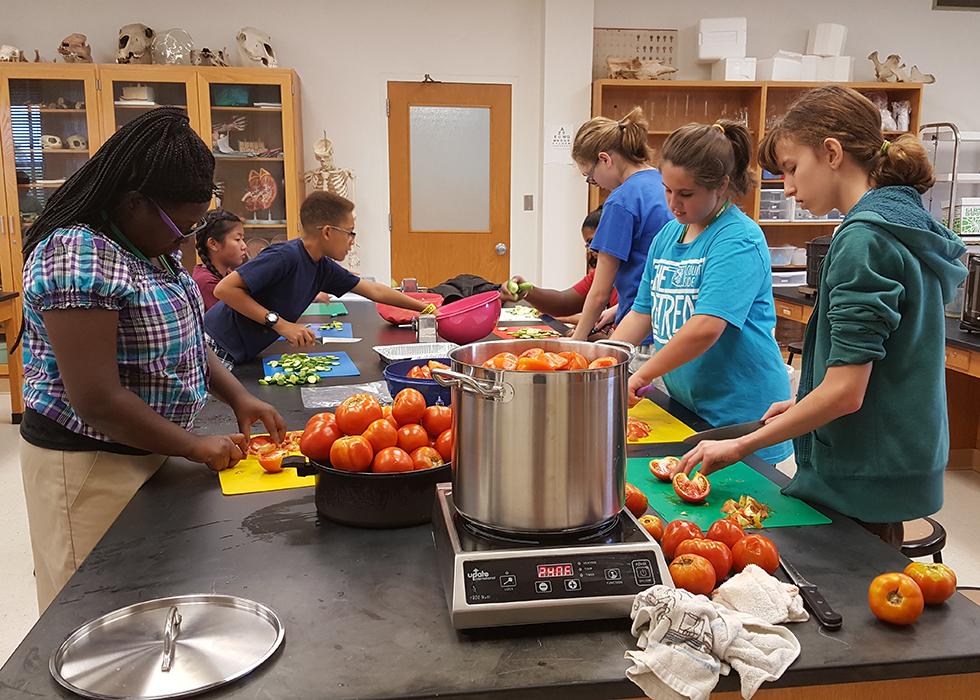 A photo of 8th and 9th grade students around a black table as they process vegetables for canning