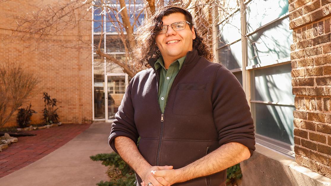 A headshot of Kalep Glandon in the courtyard of Davis Hall with trees and the building's brick walls in the background