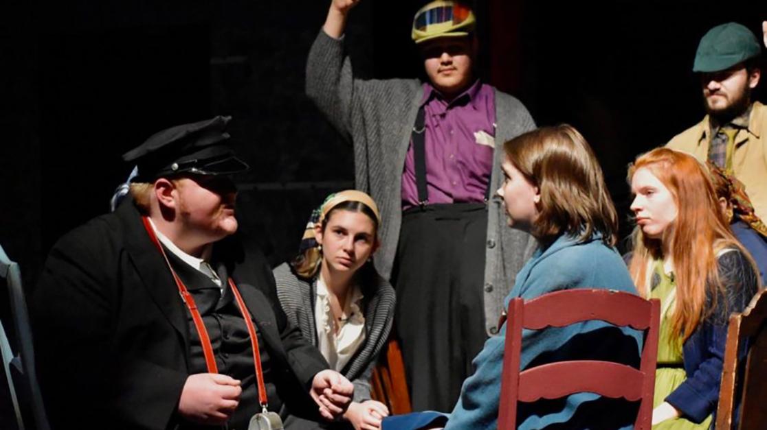 A photo of six students sitting in a rough circle in stage clothes during the dress rehearsal of A Man of No Importance