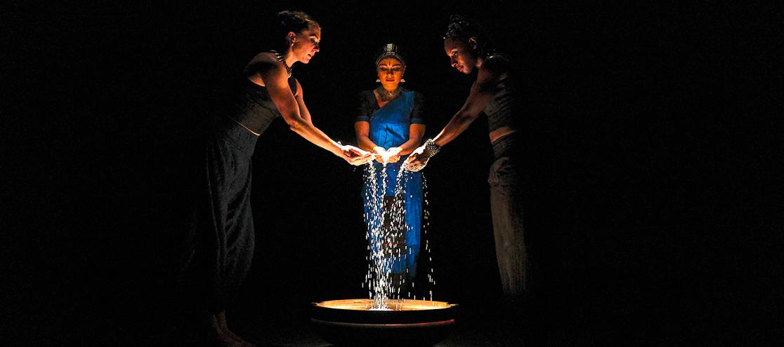 A photo of three performers on a darkened stage surrouding a brightly-lit basin while pouring grains from their hands 