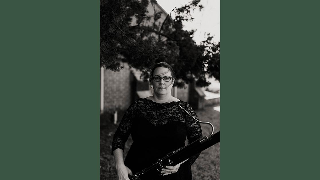 A black and white headshot of Dr. Kate Sekula holding her bassoon framed by trees in front of Davis Hall