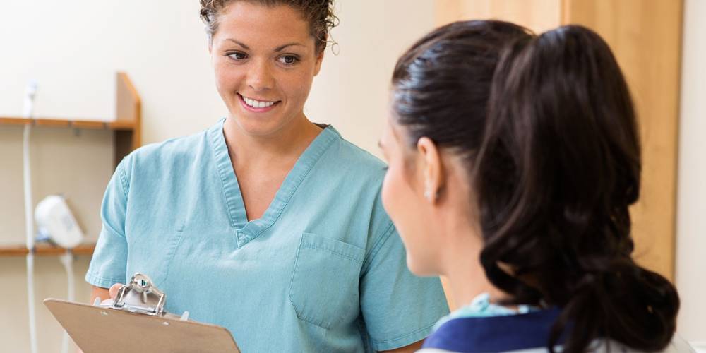 A healthcare professional in light blue scrubs with curly hair pulled back is smiling warmly while holding a clipboard and speaking with a patient. The patient, shown from behind with dark hair in a ponytail, is seated and wearing what appears to be a blue medical gown or clothing. 