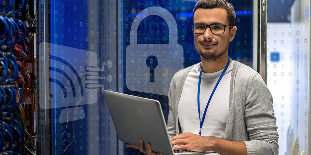 A smiling man with glasses and a beard wearing a gray cardigan over a white shirt and blue lanyard stands in a data center or server room, holding a laptop. Behind him are server racks with blue LED lighting and network cables. Overlaid on the image are translucent cybersecurity icons including a padlock, WiFi symbol, and binary code patterns, suggesting themes of network security and data protection.