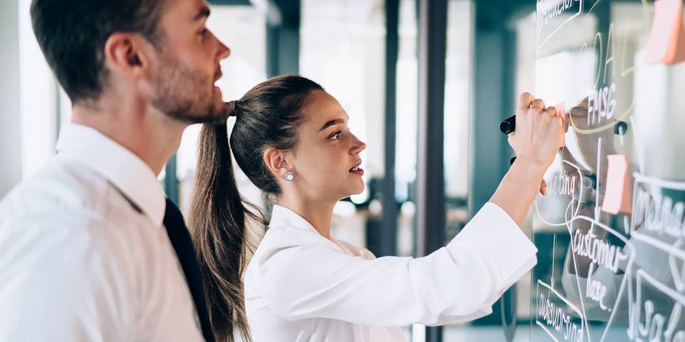 Two young professionals collaborate in a modern office, with a woman writing ideas on a transparent glass board using a black marker while a man attentively watches. The board is filled with diagrams, arrows, and sticky notes, suggesting a brainstorming or planning session.